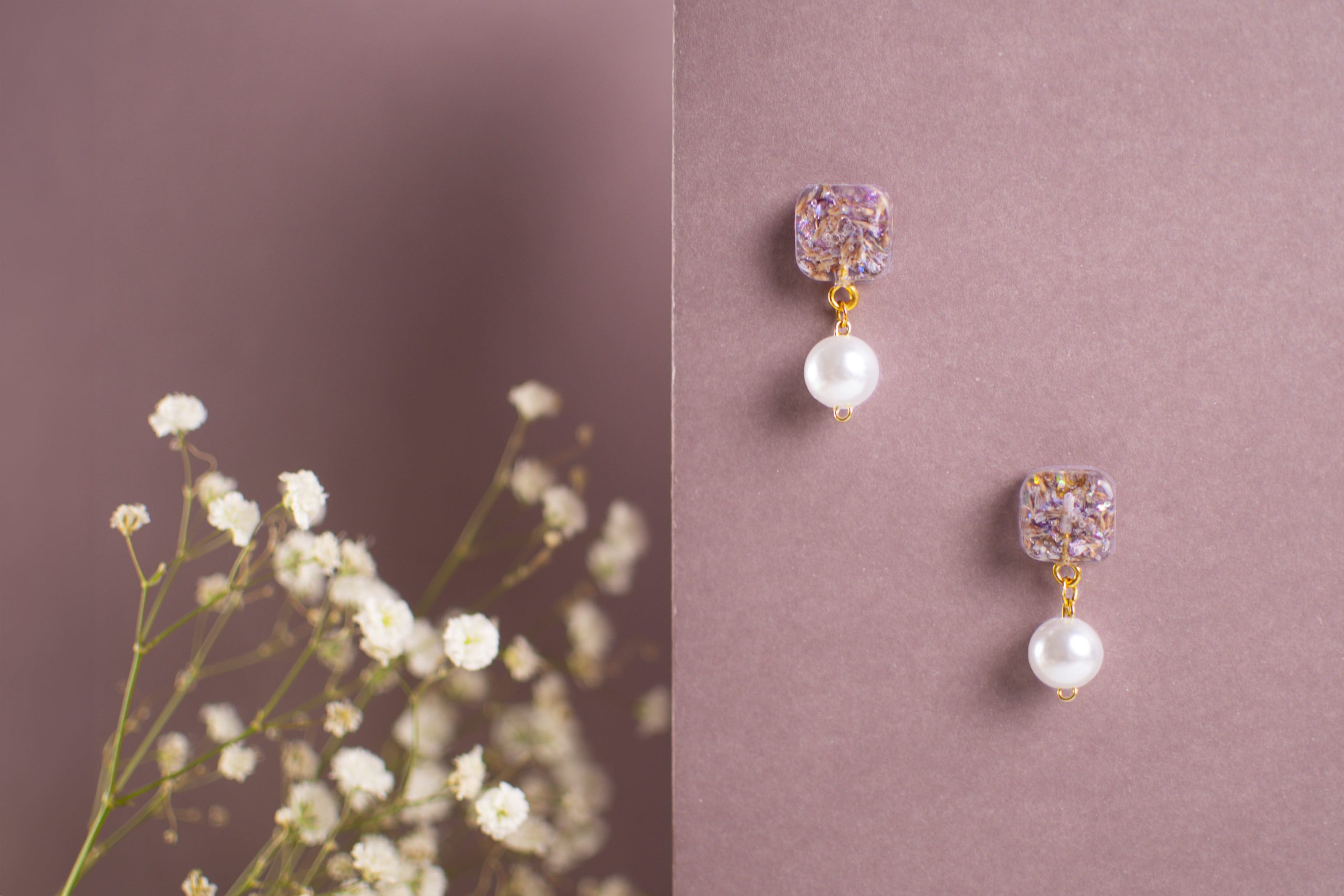 Orecchini artigianali con perla bianca pendente e base quadrata floreale viola su fondo rosa